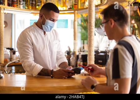 Junger lateinamerikanischer Kellner mit Gesichtsmaske, der einem Kunden per kontaktloser Bezahlung von der Bar aus verlädt. Stockfoto