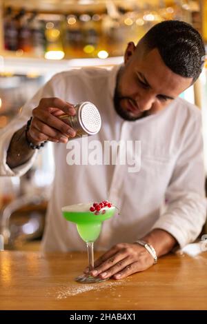 Der junge lateinamerikanische Barkeeper bereitet in einer Bar sorgfältig einen Cocktail zu. Selektiver Fokus. Stockfoto