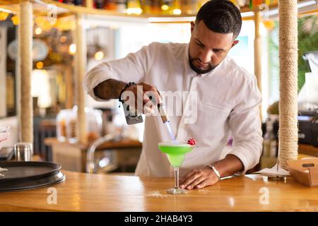 Eleganter junger lateinamerikanischer Kellner, der in einem modernen Restaurant sorgfältig ein alkoholisches Getränk zubereitete. Platz für Text. Selektiver Fokus. Stockfoto