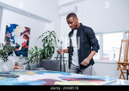 Nachdenklicher schwarzer Mann, der auf einer großen Leinwand mit Malerei auf einem Tisch schaut. Er hält einen Filzstift in der Hand. Innerhalb einer Wohnung. Stockfoto