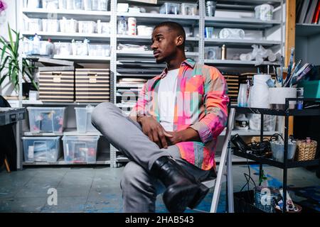 Laidback schwarzer Mann in bunten quadratischen gemusterten Hemd sitzt auf einem Stuhl in einer Werkstatt. Alle Arten von Containern auf Regalen im Hintergrund. Stockfoto