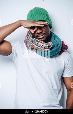 Grimacing afroamerikaner Herstellung Monocle mit seiner Hand. Er trägt einen grünen Winterhut und einen bunt gemusterten Schal. Stockfoto