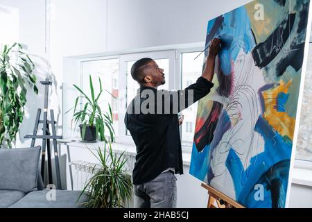 Eingetaucht schwarzer Mann Malerei auf einer großen Leinwand in seiner Wohnung. Er wendet geduldig und langsam Farbe an. Grüne Topfpflanzen und Sofa in einem Bac Stockfoto