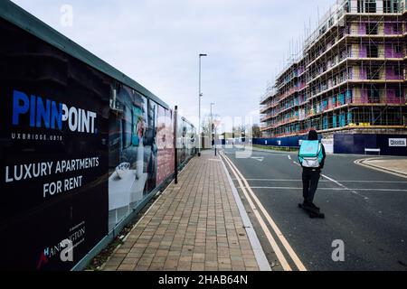 Uxbridge, Großbritannien. 9th. Dezember 2021. Ein Deliveroo-Kurier für die Lebensmittelzustellung auf einem Skateboard passiert Marketing-Hoardings außerhalb der PinnPoint-Wohnsiedlung im Londoner Stadtteil Hillingdon. PinnPoint wird als Luxus-Entwicklung vermarktet und bietet eine Reihe von Studio-, 1-, 2- und 3-Zimmer-Wohnungen zur Miete im Herzen von Uxbridge. Kredit: Mark Kerrison/Alamy Live Nachrichten Stockfoto