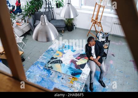 Hochwinkelbild eines schwarzen Mannes, der neben einer großen Leinwand sitzt, mit Malerei auf einem Tisch, der sie akribisch untersucht. Innerhalb einer Wohnung. Stockfoto