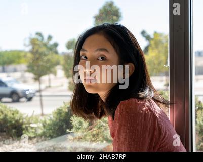 Porträt einer asiatischen Frau, die neben dem Fenster auf die Kamera schaut, horizontaler urbaner Hintergrund Stockfoto
