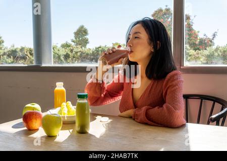 Asiatische Frau trinkt einen gesunden Obst- und Gemüse-Smoothie, sitzt neben mehr Obst und Säfte Stockfoto