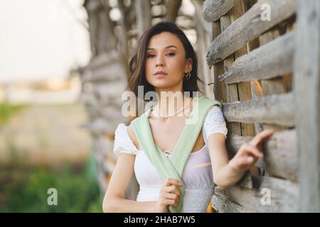 Asiatische Frau, die in der Nähe eines Tabaktrocknungsschuppens posiert und ein weißes Kleid und grüne Gummistiefel trägt. Stockfoto