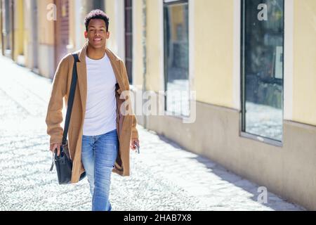 Ein junger schwarzer Mann, der die Straße entlang läuft, trägt eine Aktentasche und ein Smartphone. Stockfoto