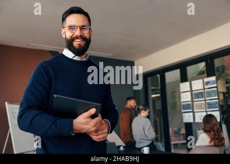 Erfolgreicher Geschäftsmann lächelt der Kamera zu, während er ein digitales Tablet hält. Glücklicher junger Geschäftsmann, der mit seinem Kollegen in einem modernen Sitzungssaal steht Stockfoto