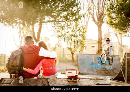 Multirassisches verliebtes Paar sitzt im Skatepark mit Musik und beobachtet Freunde auf der bmx Freestyle-Ausstellung - Urban Relationship Concept Stockfoto