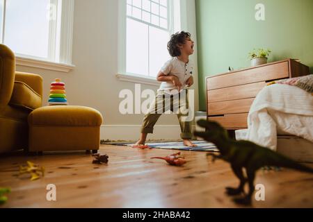 Verspielter junger Junge, der während der Spielzeit zu Hause einen T-rex-Dinosaurier imitiert. Kreativer kleiner Junge, der Spaß hat, während er in seinem Spielbereich steht. Stockfoto