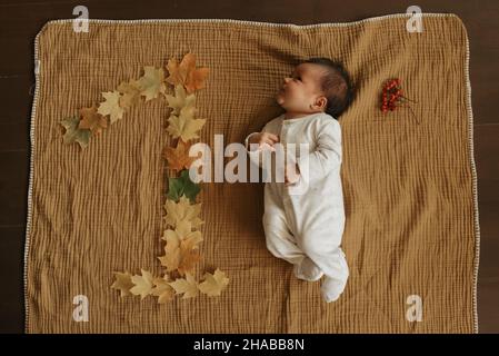 Ein Säugling liegt auf einer Musselin-Decke in der Nähe von Ahornblättern, die in der Form von 1 liegen und nach links starren. Ein niedliches Mädchen in einteiliger Kleidung ist ce Stockfoto