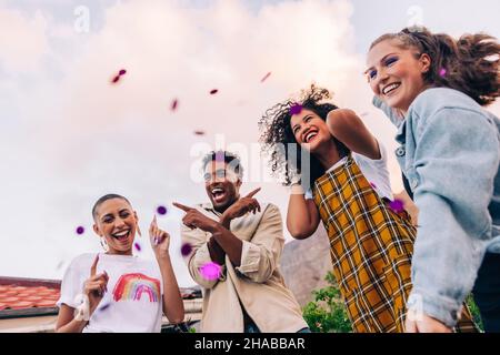 Eine Gruppe junger Leute, die während einer Party auf dem Dach tanzen. Vier glückliche Freunde feiern unter fallenden Konfetti. Multikulturelle Freunde lachen und Stockfoto