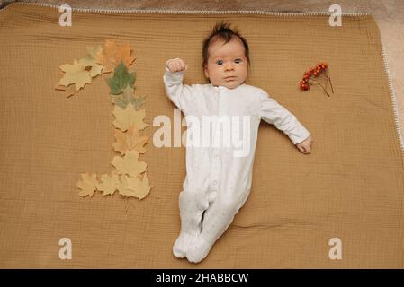 Ein eleganter Säugling liegt auf einer Musselin-Decke in der Nähe von Ahornblättern, die in Form von 1 liegen. Ein niedliches Mädchen in einteiliger Kleidung feiert ihr 1 Stockfoto