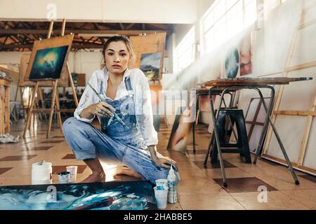 Einfallsreiche Malerin, die in ihrem Kunststudio die Kamera anschaut. Eine talentierte Künstlerin, die ein blaues Gemälde für ihr neues Kunstprojekt macht. Kreative junge Frau Stockfoto