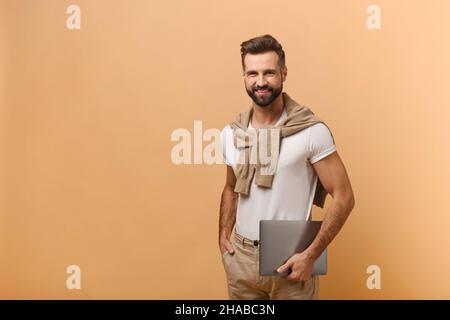 Fröhlicher Mann mit Bart, der den Laptop hält und mit einem Lächeln auf die Kamera blickt. Innenaufnahme des Studios isoliert auf orangefarbenem Hintergrund Stockfoto