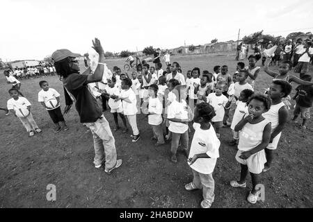 JOHANNESBURG, SÜDAFRIKA - 12. Aug 2021: Eine Graustufenaufnahme des Volunteer Soccer-Trainers, der mit jungen afrikanischen Kindern auf dem Schulhof arbeitet. Stockfoto