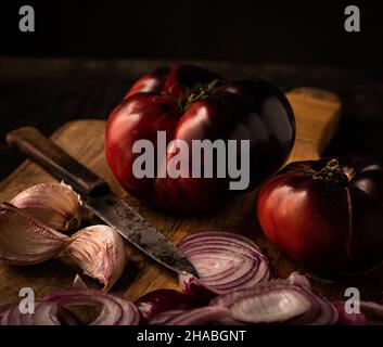 Stillleben von blauer Rindertomate, auf einem hölzernen Schneidebrett und etwas violettem Knoblauch und roter Zwiebel und einem Messer Stockfoto