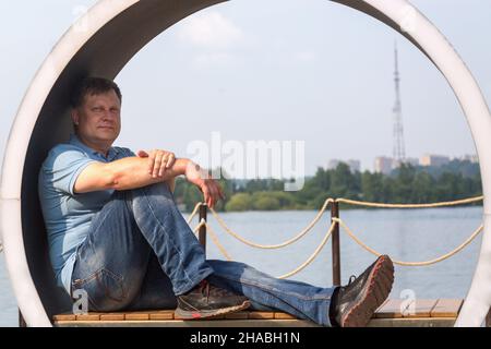 Ein erwachsener Mann sitzt in Form eines Kreises auf einer kreativen Bank und blickt auf die ruhige Oberfläche des Flusses. Stockfoto