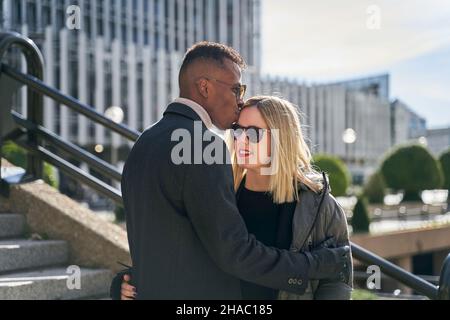 Schwarzer Mann in Sonnenbrille und Mantel umarmt und küsst die blonde Freundin auf der Stirn, während er an sonnigen Tagen in der modernen Stadt auf Treppen steht Stockfoto