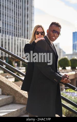 Blonde Frau in Sonnenbrille, die an einem sonnigen Tag auf der Straße hinter einem afroamerikanischen Freund im trendigen Mantel auf der Treppe steht Stockfoto