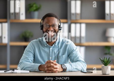 Lächelnder, tausendjähriger afroamerikanischer bärtiger Kerl in Brille und Headset, der am Laptop arbeitete und auf die Kamera schaute Stockfoto