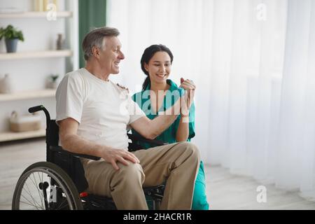 Junge Physiotherapeutin hilft älteren Mann im Rollstuhl mit Kurzhanteln zu trainieren, um sich von Verletzungen zu erholen Stockfoto