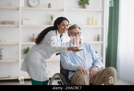 Junge Krankenschwester mit Fernbedienung, die Kanäle im Fernsehen für einen älteren Mann im Rollstuhl im Altersheim umschaltet Stockfoto