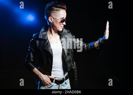 Stilvolle kurzhaarige Brünette Frau in schwarzer Lederjacke trägt Sonnenbrille macht Stop-Geste mit der Hand auf schwarzem Hintergrund mit blauem Licht. Verwirrtes und negatives Konzept. Stockfoto