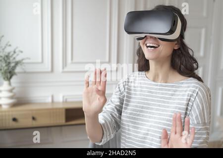 Junge, aufgeregte Frau, die ein VR-Headset trägt, interagiert mit Augmented World und spielt zu Hause Spiele Stockfoto