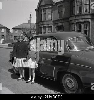 1958, historische, zwei junge Mädchen, Schwestern, stehen draußen in einer Straße neben dem Auto ihres Vaters, einem französischen Renault Dauphine, einem 4-türigen Economy-Auto mit Heckmotor, einem Nachfolger des Renault 4CV. Zwischen 1957 und 1961 wurden Dauphines im Renault-Werk Acton in West-London montiert und einer wurde sogar von HM the Queen gefahren. Ein Aufkleber für das Fahrzeug befindet sich auf der hinteren Scheibe. Das Fahrzeug gewann die Rallye Monte Carlo 1958 und war ein weltweiter Erfolg für Renault. Stockfoto