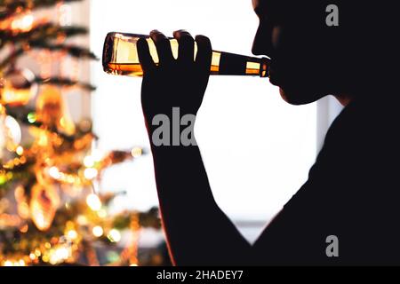 Weihnachts- und Alkoholproblem. Einsamer Mann, der Bier trinkt. Weihnachtsbaum. Alkoholismus allein während des Urlaubs. Trinken Sie aus einer Flasche Bier. Trauriger Typ mit Depressionen. Stockfoto