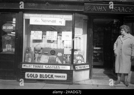 1958, eine Dame, die in der Tür eines Tabakwarenhandels steht, John Isaacs, Half Moon Street, Sherborne, Dorset, England, Großbritannien. John Isaacs war ursprünglich Friseur und die Räumlichkeiten zu einer Zeit, kombiniert mit dem Verkauf von Tabak. Im Fenster, Werbung für Peter Stuyvesant Zigaretten, "der internationale Pass zum Rauchvergnügen" und "rund um den Globus, können Sie sicher auf Peter Stuyvesant spielen." Die Marke wurde ‘in Deutschland gegründet und mit ihrer „amerikanischen Mischung“ aus Tabak aus verschiedenen Ländern als international propagiert. Außerdem wurden Wills Three Castles Cigarettes, Gold Block & Gold Flake gezeigt. Stockfoto