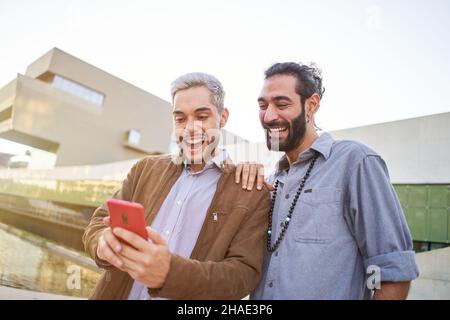 Lächelndes schwule Paar zusammen mit Telefonen. Glückliche Menschen, die etwas auf dem Handy beobachten. Stockfoto