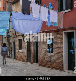 Eine junge Frau geht durch eine enge Gasse in Venedig. Wäsche hängt zum Trocknen auf der Wäsche über ihr. Blaue Partyballons hängen an Fenstern und Türen. Stockfoto