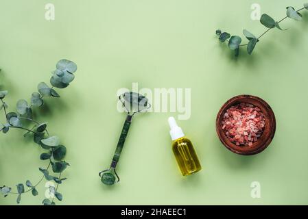 Kosmetisches Serum in Glasflasche, rosa himalaya-Salz, Gesichtswalze und Eukalyptusblätter auf hellgrünem Hintergrund. Hautpflege, natürliche Bio-Kosmetik Stockfoto