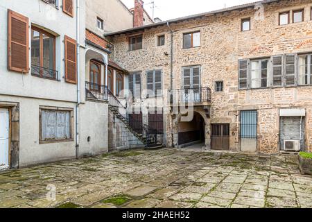 Eine schmiedeeiserne Außentreppe führt zu einer Galerie in einem Innenhof in der Altstadt von Saint Chamond, Département Loire, Frankreich. Schuttsteinmauern, Holzläden und ein rundbogiger Durchgang kennzeichnen den moosbewachsenen Innenhof. Stockfoto
