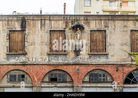 Die verwitterte Fassade eines ehemaligen Gaswerks im alten Stadtkern von Saint Chamond trägt noch Spuren der Aufschrift "Société du Gaz de St-Chamond", Departement Loire, Frankreich. In einer kleinen Nische über dem ehemaligen Firmenschild steht eine Statue eines Heiligen, und rostige Rollläden bedecken die Fenster. Im Erdgeschoss ist auf den Ziegelbögen das Wort "Magasin" (Laden) zu lesen. Stockfoto