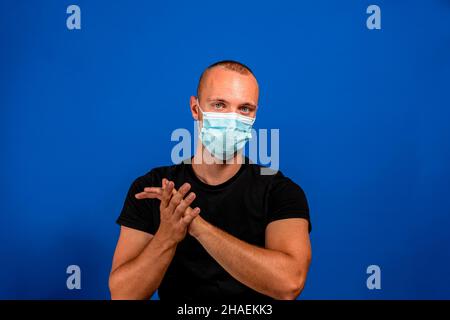 Der junge kaukasische Mann, der in einem schwarzen T-Shirt gekleidet ist und die Toilettenverwaltung applaudiert, trägt eine Maske, um das Coronavirus zu verhindern. Isoliert auf blau Stockfoto