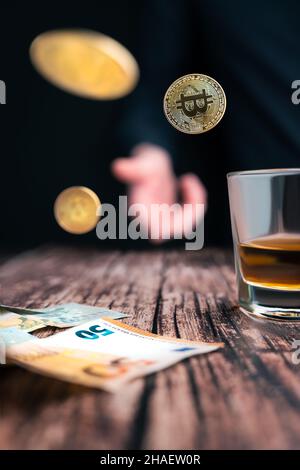 Stillleben mit drei goldenen Bitcoins, die über den Holztisch geworfen wurden, mit einem Glas Whiskey und Euro-Banknoten darauf. Spielkonzept. Kryptowährung Stockfoto