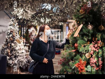 Junge Frau in Gesichtsmaske macht Neujahr Shopping auf saisonale Verkäufe während Pandemie, zu Fuß zwischen Reihen von Weihnachten festliche Dekorationen, künstliche Weihnachtsbäume, glänzende Spielzeug und bunte Girlanden Stockfoto