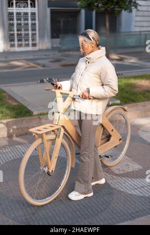 Hoher Winkel der erwachsenen Frau in stilvoller Oberbekleidung mit Smartphone, während auf dem Bürgersteig in der Nähe von Holz Öko-Fahrrad stehen. Stockfoto