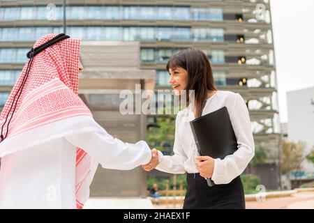 Eine junge, angenehme kaukasische Geschäftsfrau begrüßt den männlichen arabischen Kunden bei einem Treffen im Freien, zwei lächelnde Geschäftspartner, saudi-Mann und eine europäische Frau, die sich nach einem erfolgreichen Deal die Hände schüttelten Stockfoto