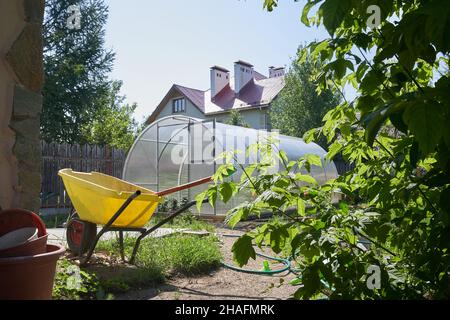 Ein modernes Gewächshaus aus Polycarbonat. Landwirtschaftliche Werkzeuge und eine Katze mit einem Rad im Garten. Das Konzept des modernen Gartenbaus. Hochwertige Fotos Stockfoto