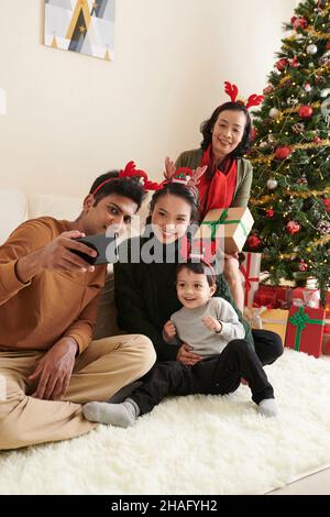 Glücklicher junger Mann, der am geschmückten Weihnachtsbaum Selfie mit Frau, Sohn und Schwiegermutter macht Stockfoto