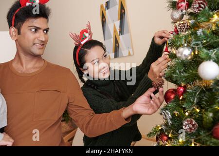 Lächelndes junges Paar, das zu Hause Dekorationen am Weihnachtsbaum hängt, wenn es sich auf die Feier vorbereitet Stockfoto