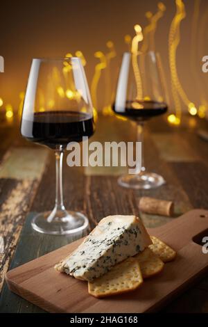 Stillleben von zwei Gläsern Wein, Käse mit blauem Schimmel und Crackern auf einem Holzbrett vor dem Hintergrund einer Neujahrskirlande. Vorderansicht. Stockfoto
