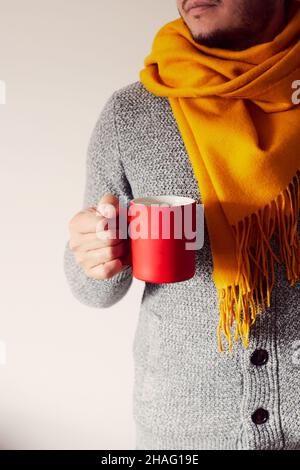 Ein Mann mit einer roten Tasse Kaffee in der Hand, der eine Strickjacke und einen Schal trägt. Das Konzept des Herbstes. Vorderansicht. Stockfoto