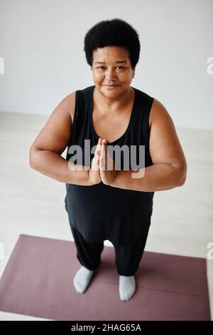 Vertikales Hochwinkelporträt einer schwarzen älteren Frau, die drinnen Yoga genießt und die Kamera anlächelt Stockfoto
